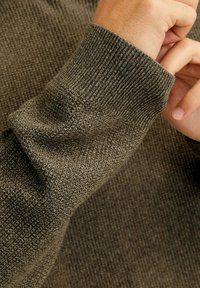 Tissu en tricot vert avec une surface texturée. Présente des poignets côtelés et une apparence douce, mettant en valeur un motif de tissage serré.