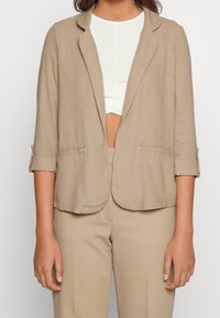 Beżowa lniana marynarka z klapami z wycięciem, podwiniętymi rękawami i dwoma przednimi kieszeniami, zestawiona z białą bluzką i dopasowanymi beżowymi spodniami.