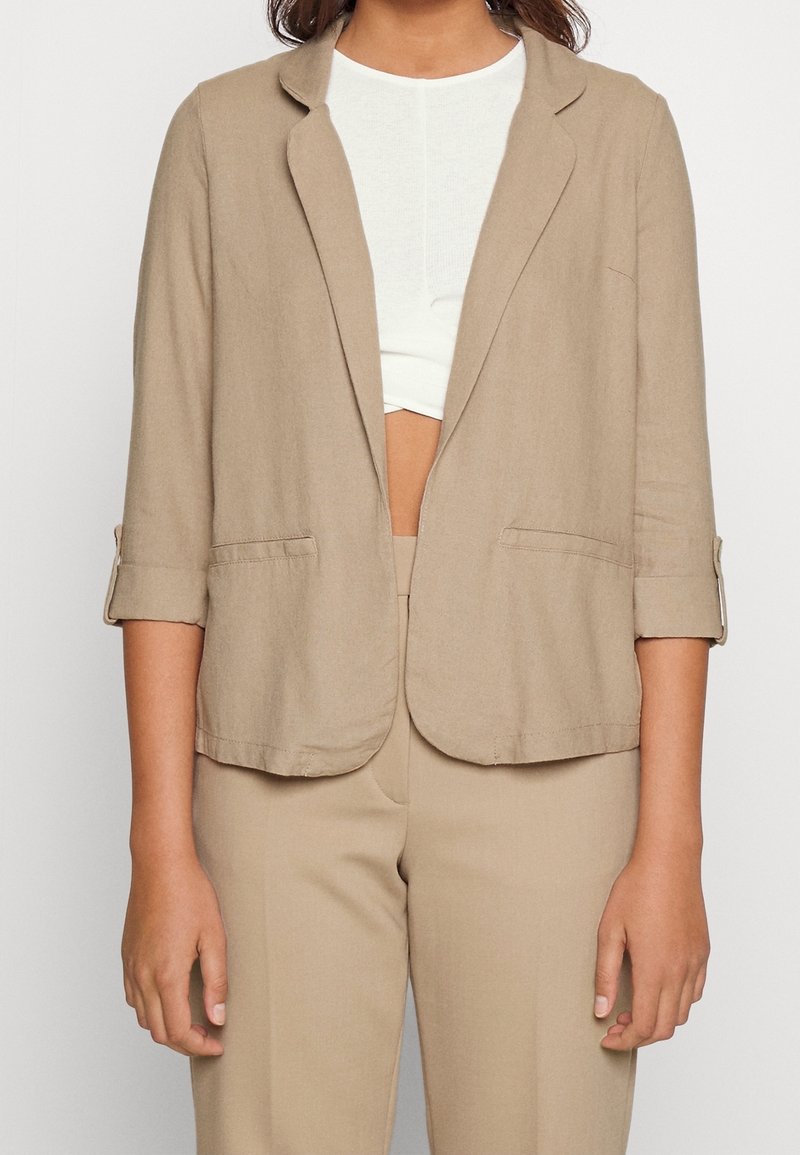 Beżowa lniana marynarka z klapami z wycięciem, podwiniętymi rękawami i dwoma przednimi kieszeniami, zestawiona z białą bluzką i dopasowanymi beżowymi spodniami.
