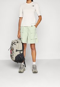 Světlé béžové tričko s krátkým rukávem a černým logem, v kombinaci s mátově zelenými cargo kraťasy, šedým batohem s více kapsami a šedými trekingovými botami.