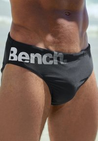 Crne kupaće gaće s teksturiranim elastičnim pojasom na kojem je u bijeloj boji natpis "Bench." Glatka tkanina s prilagođenim dizajnom.