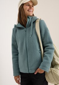 Veste en polaire de couleur turquoise muet avec fermeture éclair frontale, deux poches latérales et une capuche. Doublure intérieure lisse et surface extérieure texturée.