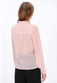 Blusa rosa chiaro con colletto arricciato, tessuto morbido e dettagli metallici. Maniche lunghe con polsini con bottoni, tessuto trasparente.
