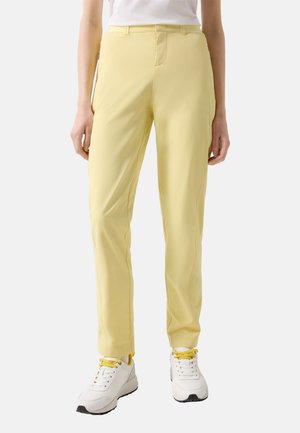 Osoba ubrana w dopasowane jasnofioletowe spodnie z białymi trampkami z żółtymi sznurówkami, stojąca na jednolitym tle.