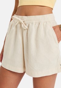 Shorts en lin beige clair avec une taille élastique, fermeture à cordon, et poches latérales. Le tissu a une finition légèrement texturée.