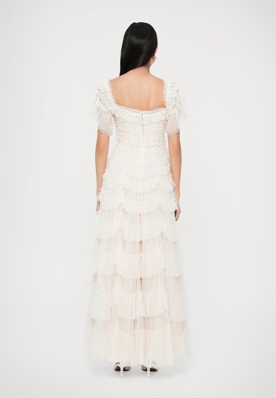 Dlouhé bílé šaty s vrstvenými volánky, průhlednými balónovými rukávy a čtvercovým výstřihem. Obsahují zip na zadní straně a texturovanou tkaninu.