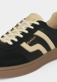 Baskets GANT noires et beiges avec lacets blancs, semelle en caoutchouc et détail du logo sur le côté et la languette.