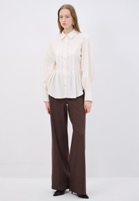 Crème blouse met lange mouwen, knoopsluiting, geplisseerd ontwerp en een gestructureerde kraag, gecombineerd met hoge bruine wijde broek.