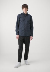 Marineblå skjorte med hvitt sikksakkmønster, kombinert med mørke bukser og hvite joggesko. Skjorten har klassisk krage og lomme.