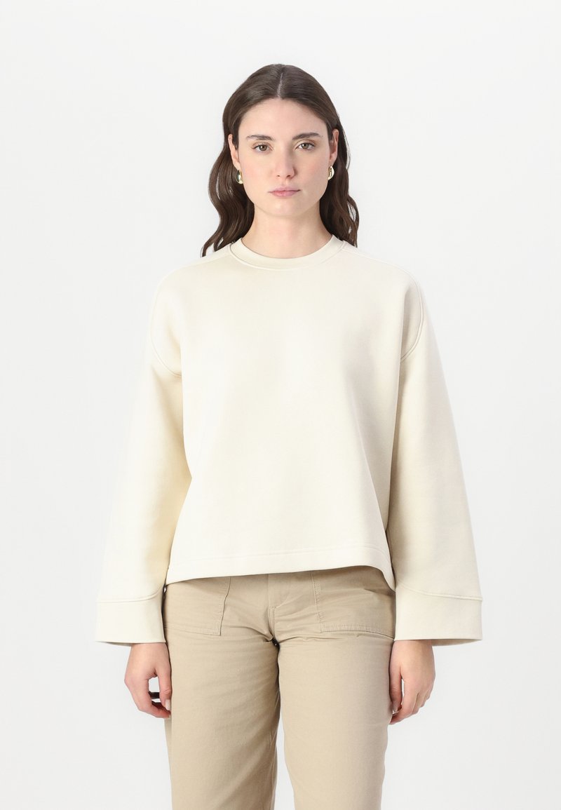 Felpa color crema con scollatura rotonda, spalle scese e polsini larghi. Il tessuto appare morbido con una texture liscia. Indossata con pantaloni beige.