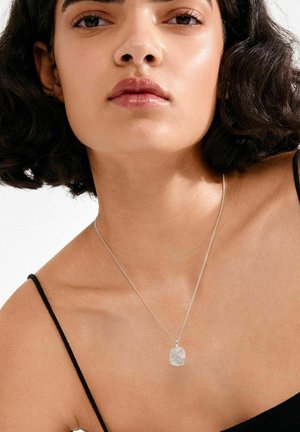 Zilveren ketting met een textuurrijk, ovaal hangertje, gedragen op blote huid, met een zwart topje met dunne bandjes. Glans- en reflectieaccenten zichtbaar.