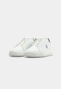 Une paire de baskets basses blanches avec des lacets, semelles épaisses et le logo "CK" de Calvin Klein sur les côtés extérieurs.