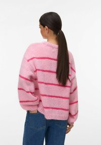 Pull en tricot rose avec de larges rayures roses et magenta, présentant un design ample, des épaules tombantes et un ourlet côtelé. Associé à un jean bleu.