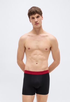 Νέος άνδρας που στέκεται με τα χέρια πίσω από την πλάτη, φοράει μαύρα boxer briefs με κόκκινη μέση, σε απλό λευκό φόντο.