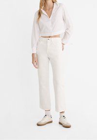 Bílá zkrácená košile na knoflíky spojená s krémovými džíny s rovnými nohavicemi a béžovými teniskami, vyznačující se čistým a minimalistickým designem.