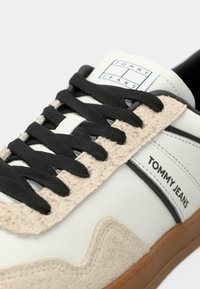White sneaker with black laces, featuring a textured beige suede panel and a rubber gum sole. "TOMMY JEANS" logo on the side.