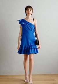 Robe bleue à une épaule avec un ourlet volanté, tissu léger et accent floral sur l'épaule. Portée avec un petit sac noir et des sandales à lanières.