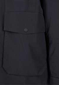 Čierna látka s textúrovaným štruktúrovaným vzorom; obsahuje prednú vrecko s klopou a okrúhlym gombíkovým uzáverom.