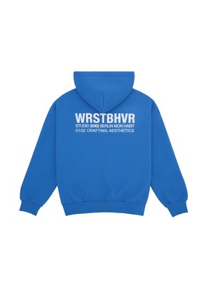 Blå hættetrøje med hvid tekst på ryggen: "WRSTBHVR STUDIO (030) BERLIN MON HABIT 0102 CRAFTING AESTHETICS," ensfarvet baggrund.