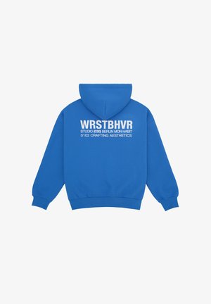 Blå hættetrøje med hvid tekst på ryggen: "WRSTBHVR STUDIO (030) BERLIN MON HABIT 0102 CRAFTING AESTHETICS," ensfarvet baggrund.