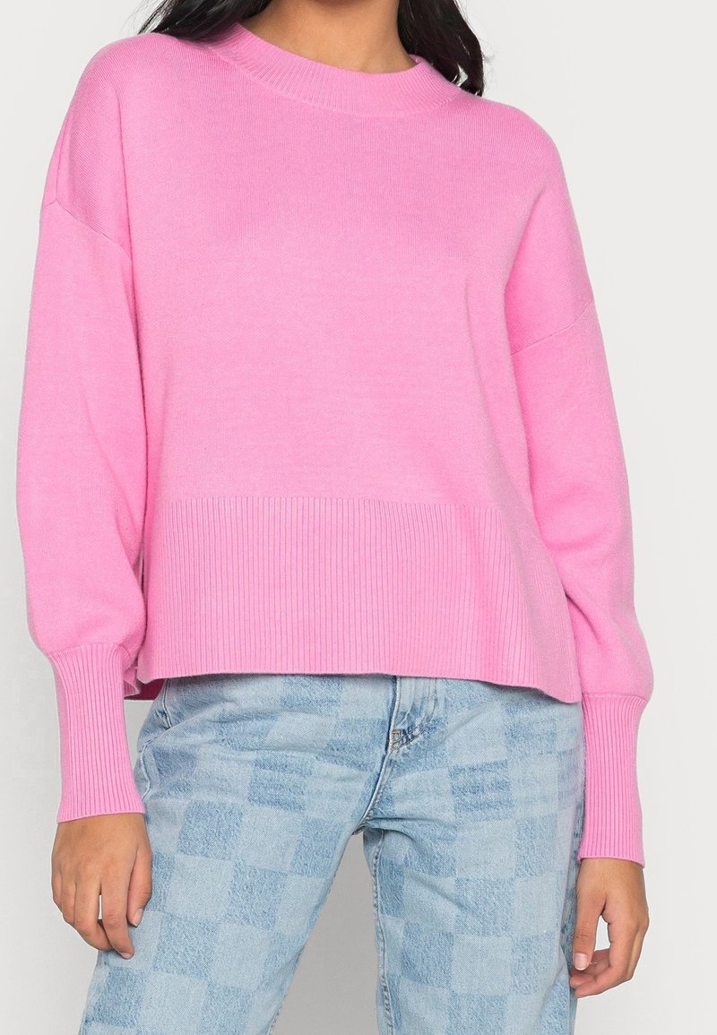 Różowy sweter na drutach z prążkowanym dekoltem i krawędzią. Ma luźne rękawy i krótki fason, w parze z jasnoniebieskimi, kratkowymi dżinsami.