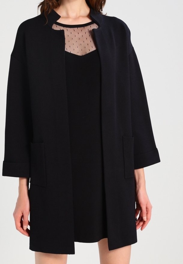 Femme portant un manteau noir mi-long sur une robe noire avec un décolleté transparent à pois, debout devant un fond uni.