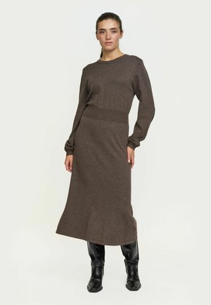 Mujer con vestido midi marrón de manga larga y cintura ajustada, combinada con botas negras hasta la rodilla, de pie contra un fondo claro.
