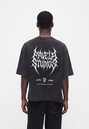 Άτομο που φοράει μαύρο φαρδύ t-shirt με λευκό κείμενο σε φυλετικό στιλ "Savelah Studios" και γραφικά στην πλάτη, συνδυασμένο με μαύρο παντελόνι.