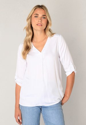 Vrouw met lang blond haar die een witte V-hals blouse met opgerolde mouwen en lichtblauwe spijkerbroek draagt tegen een effen achtergrond.