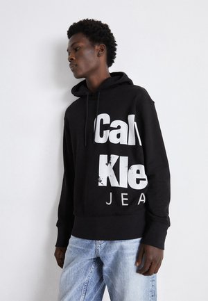 Felpa nera con grande scritta bianca "Calvin Klein Jeans". Presenta un cappuccio con coulisse e polsini a coste, abbinata a jeans di denim blu chiaro.