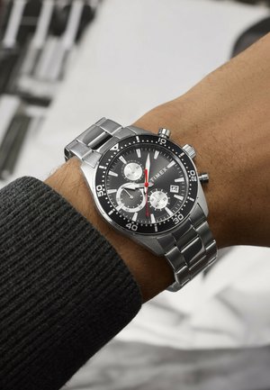Håndled med et sølvfarvet Timex kronografur med sort urskive, tre underskiver, datovindue og rustfrit stål armbånd.