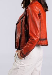 Rode leren motorjack met asymmetrische rits, gematteerde afwerking en opvallende naden. Gecombineerd met lichte broek, zijaanzicht getoond.