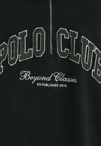 Svart tröja med kvartdragkedja, med "POLO CLUB" i upphöjd grå brodyr och "Beyond Classics" i vit skrivstil, grundad 2012.