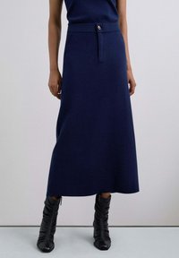 Vrouw in een marineblauwe midi-rok met een gouden sluiting en zwarte enkellaarzen met hakken, staand tegen een effen lichte achtergrond.