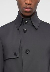 Ciemnoszary płaszcz trencz z klapowanym kołnierzem, dwoma czarnymi guzikami i detalem kieszeni z klapą. Gładka faktura tkaniny, prosty krój.