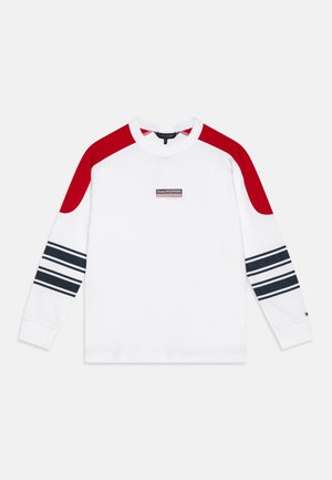 Biała koszula z długim rękawem z czerwonymi panelami na ramionach, czarno-białymi paskami na rękawach oraz małym prostokątnym logo Tommy Hilfiger na piersi.
