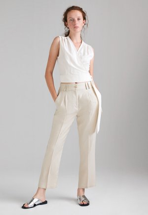 Donna con capelli ricci che indossa un top bianco smanicato a fascia, pantaloni beige a vita alta e sandali argento a ciabatta su uno sfondo grigio neutro.