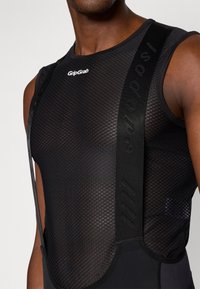 Capa base sin mangas de malla negra con logo; cuenta con correas elásticas y tejido texturizado, diseñado para la transpirabilidad y la comodidad durante su uso.