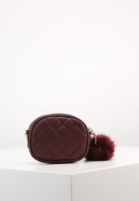 Burgundy quilted handbag with a rounded shape, gold chain strap, and a fluffy pom-pom accent. Smooth texture with defined stitching patterns.