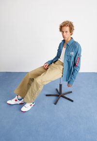 Svetlomodrá košeľa na gombíky, béžové široké nohavice s vreckom, biele tenisky s červenými a modrými akcentmi. Sedí na čiernej stoličke.