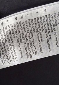 Etichetta di cura del tessile. Tessuto bianco con testo nero, che elenca i materiali: 70% cotone, 30% poliestere. Include più lingue. Layout piatto.