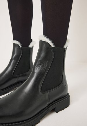 Botas de tobillo de cuero negro con paneles laterales elásticos, que cuentan con un suave borde de pelo sintético blanco en la parte superior y una suela de goma gruesa.