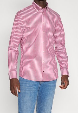 Mand iført pink og hvid lodretstribet skjorte med knapper og blå jeans, stående foran en enkel lys baggrund.