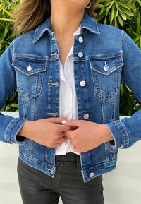 Denim jacket in donkerblauw met een getailleerd ontwerp, zilveren knopen, borstzakken en lichte slijtage op de schouders. Gecombineerd met een wit shirt.