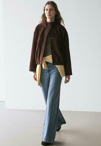 Braune Wildlederjacke über einem gestreiften Hemd, mit einer gelben Krawatte in der Taille, kombiniert mit hellblauen Schlagjeans und schwarzen Schuhen.