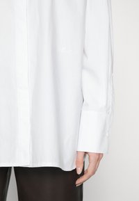 Camicia bianca a maniche lunghe con polsini abbottonati indossata con pantaloni neri, mano rilassata lungo il fianco, testo ricamato sottile nell'area del petto.