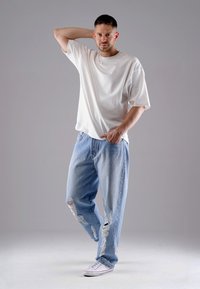 Weißes, übergroßes T-Shirt, lässig geschnittene hellblaue, ausgefranste Jeans mit Rissen und weiße Sneaker mit roten Akzenten vor einem neutralen Hintergrund.