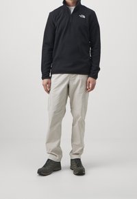Fleece mikina v černé barvě s čtvrtinovým zipem, v kombinaci se světle béžovými cargo kalhotami a tmavými turistickými botami. Minimalistický design a praktické detaily.