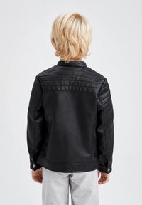 Černá koženka s strukturovaným designem, s prošívanými detaily na ramenech a zapínáním na knoflík, nošena se šedými kalhotami.