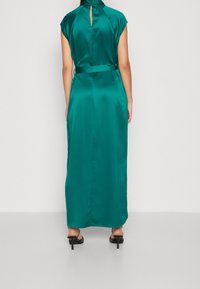 Lång grön satinklänning med hög krage, korta ärmar och ett nyckelhål baktill. Knytning i midjan. Slät textur.